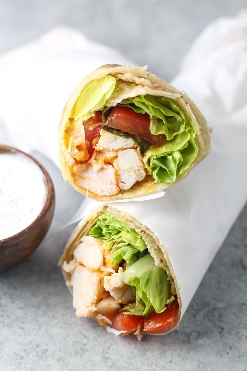 Two halves of a low FODMAP buffalo chicken wrap stuffed with chicken, lettuce, and tomato. A side of homemade ranch on the side.