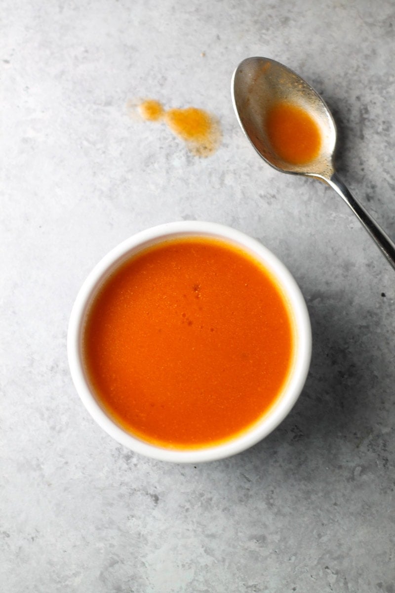 Looking down at a bowl of buffalo sauce. A spoon previously dipped in the sauce sits off to the side.