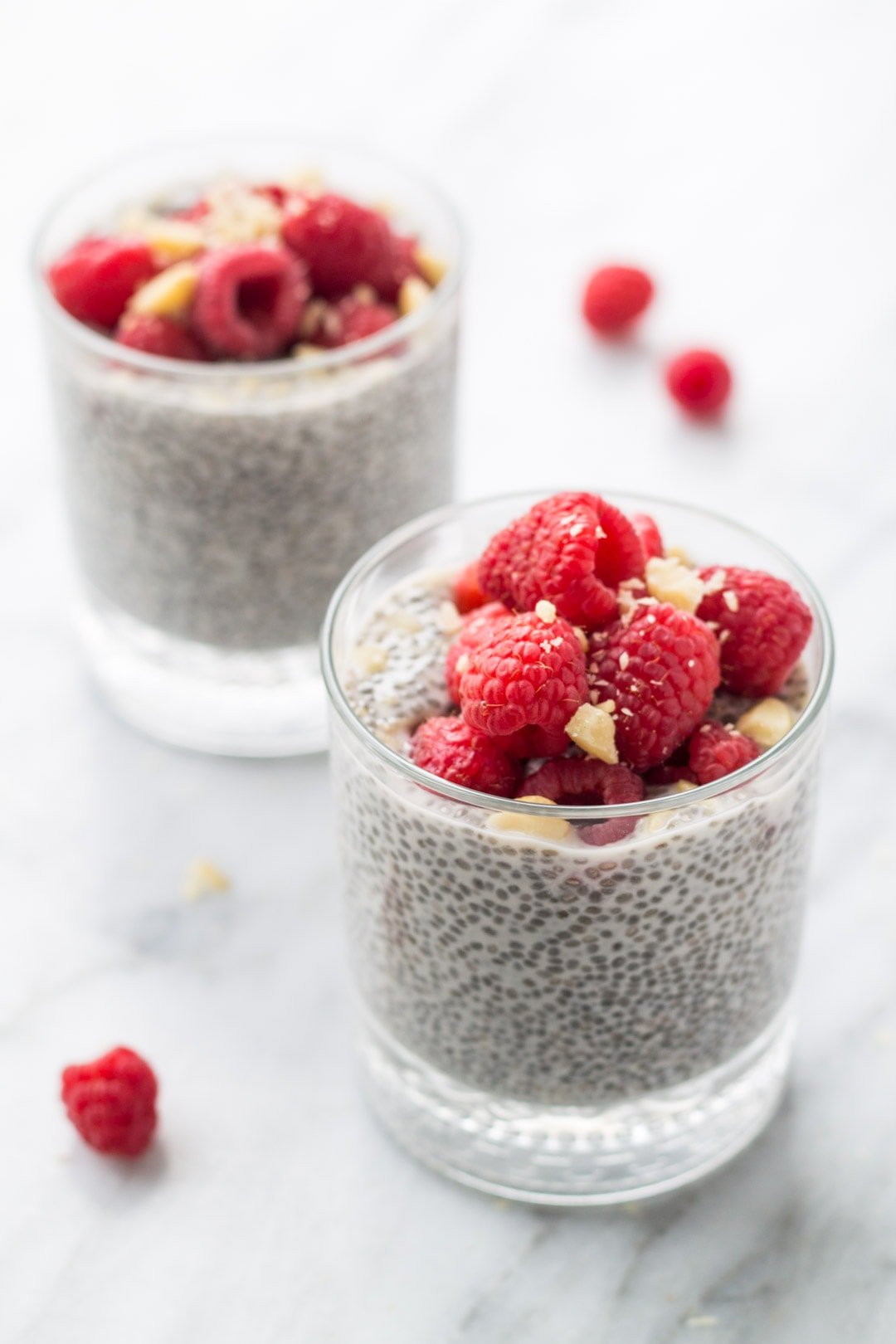 Side shot of two cups filled with vanilla chia pudding topped with fresh raspberries and macadamia nuts.