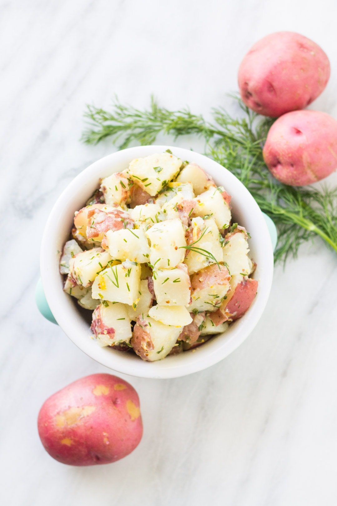 A bowl of low FODMAP dilly potato salad surrounded by several red potatoes and a sprig of fresh dillweed.