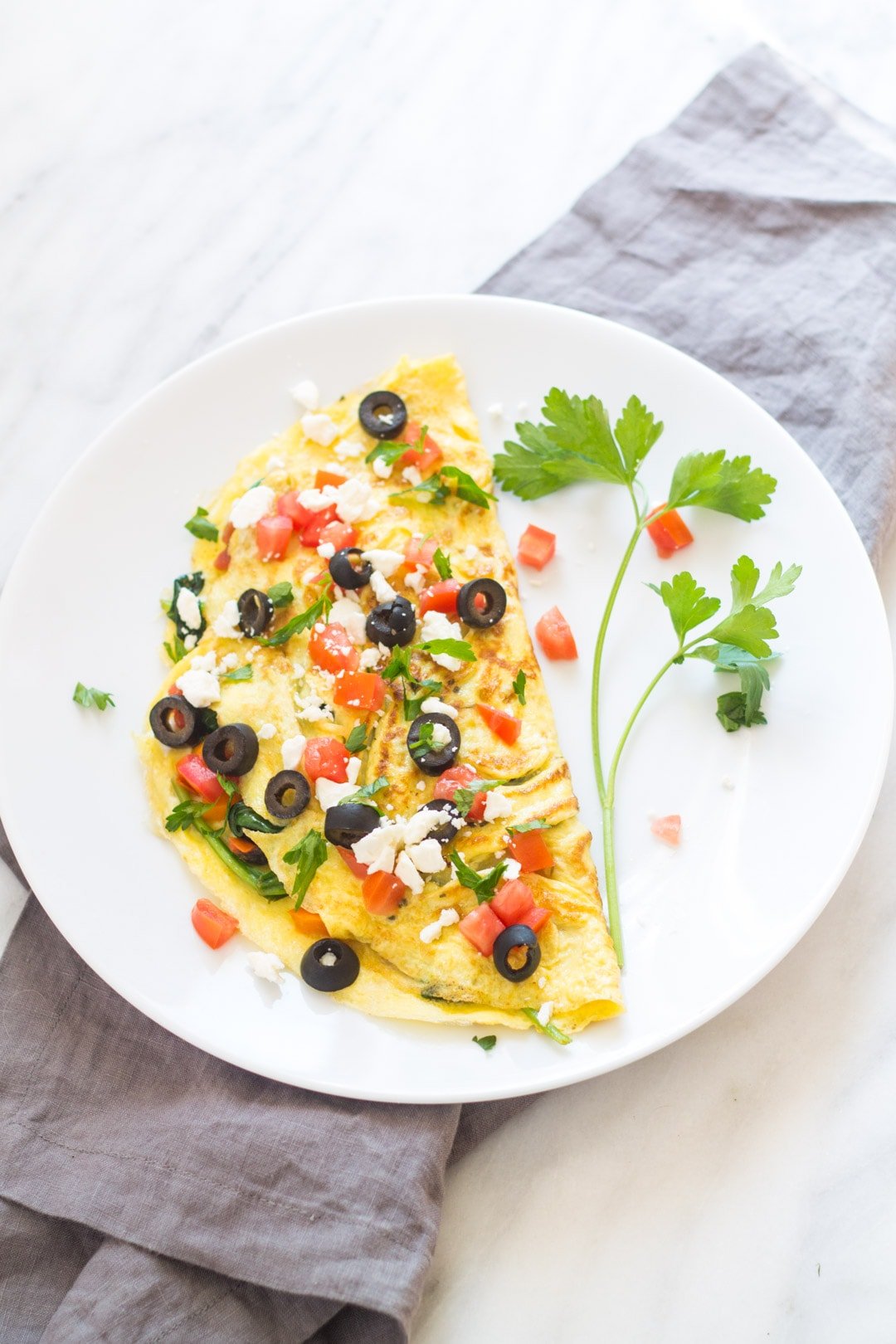 An omelet topped with tomatoes, black olives, feta cheese, and fresh parsley sit on a white plate. 