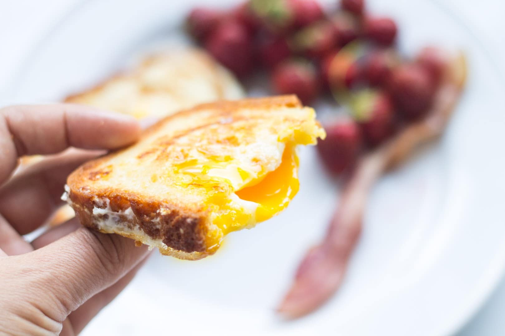 A hand holding a slice of toasted bread containing an over-medium egg cooked into the center.