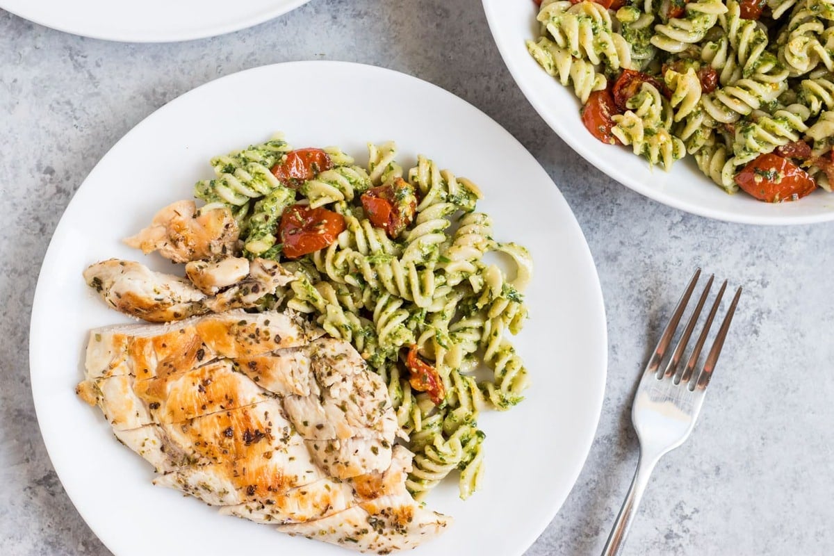 A plate and serving bowl filled with brown rice pasta tossed with homemade pesto, grilled chicken, and roasted tomatoes.