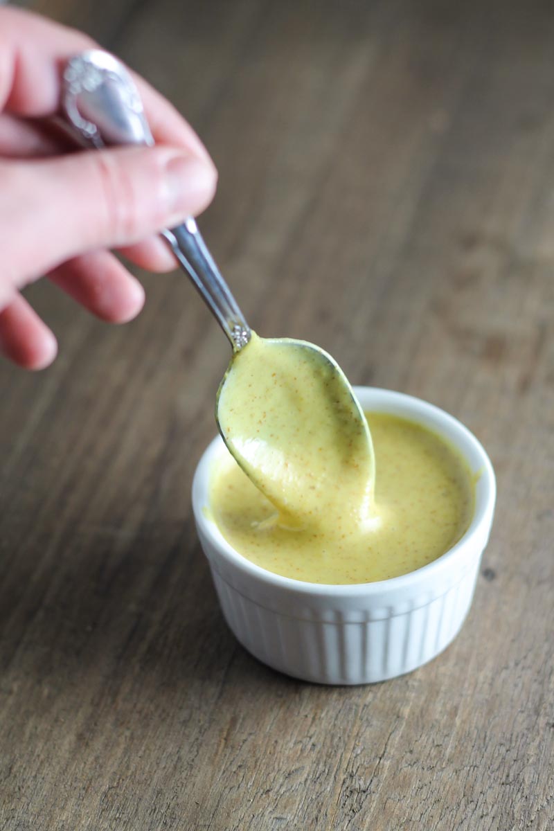A spoon dipped into a small white bowl containing creamy maple mustard