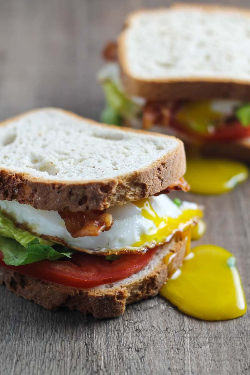 Close up of two low FODMAP sandwiches filled with bacon slices, a fried egg, tomato slices, and lettuce leaves.