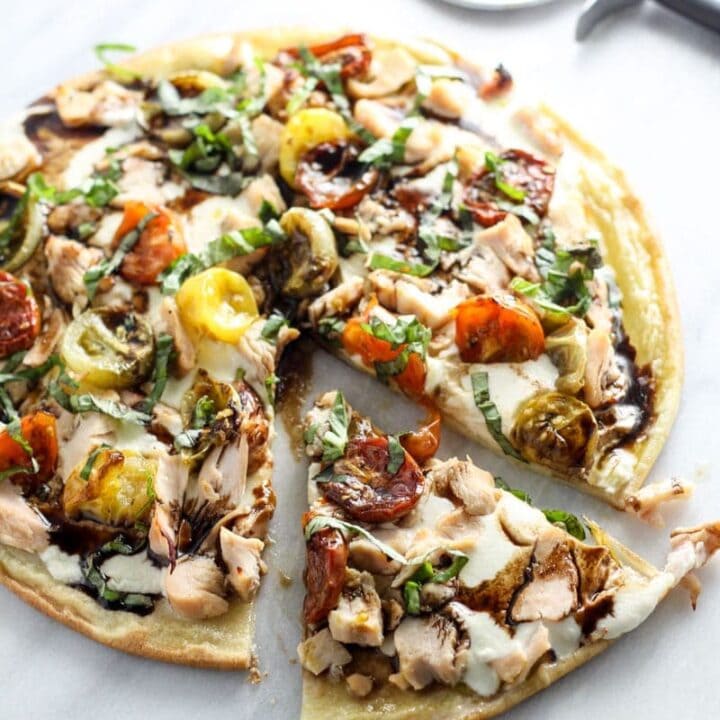 A sliced pizza topped with chicken, multi-color roasted tomatoes, fresh basil, mozzarella, and balsamic.