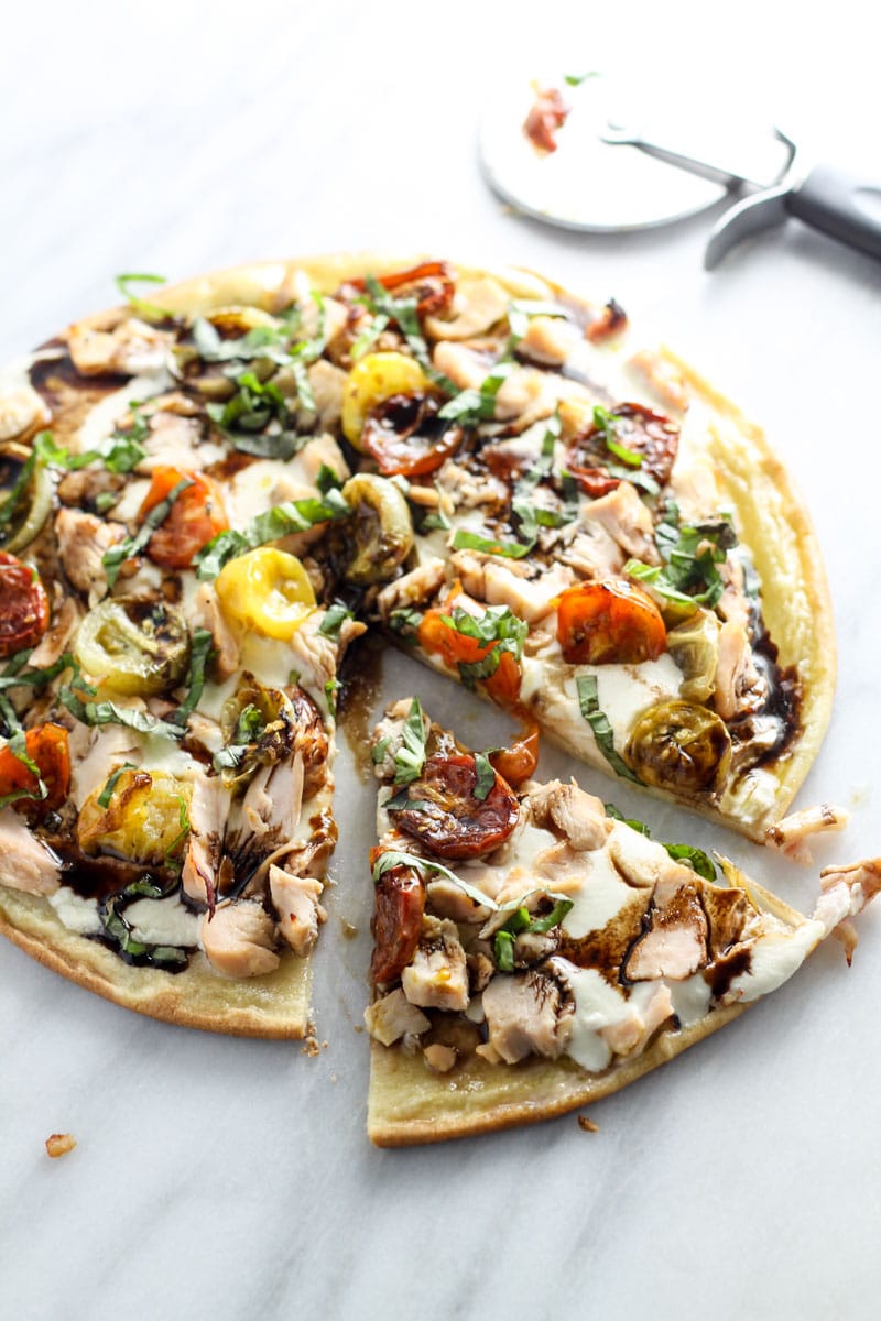 A sliced pizza topped with chicken, multi-color roasted tomatoes, fresh basil, mozzarella, and balsamic. 