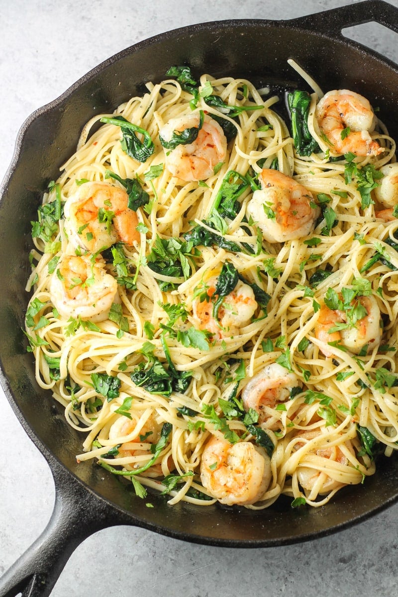 A cast-iron filled with buttery linguini, cooked shrimp, and wilted spinach topped with lots of chopped fresh parsley.