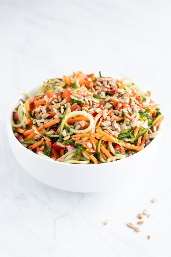 A side shot of a white bowl filled with Low FODMAP Asian Zoodle Salad. A handful of sunflower kernels are scattered in the foreground.