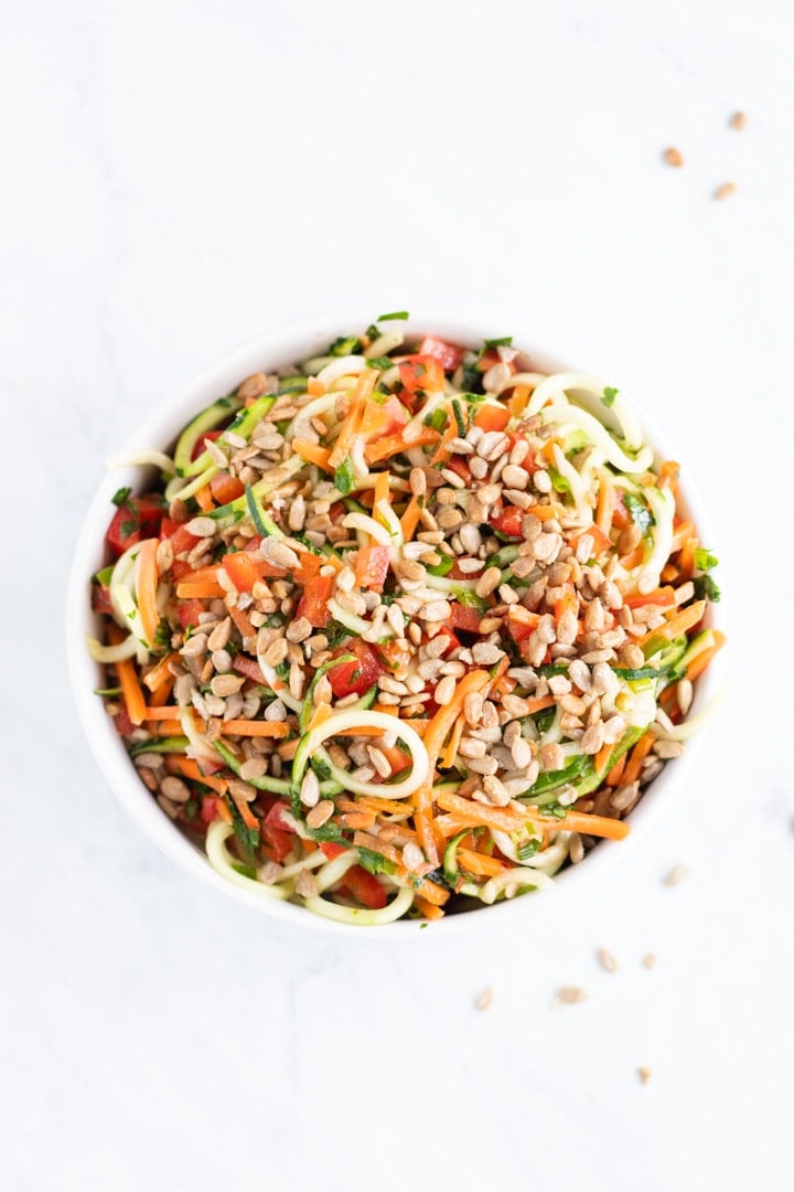 Overhead shot of a white bowl sitting on a white marble slab. The bowl is filled with low FODMAP Asian Zoodle Salad. There are random sunflower kernels artistically scattered around the bowl.