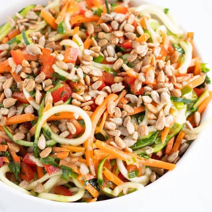 A close up of a white bowl filled with low FODMAP Asian Zoodle Salad: zucchini noodles, carrot shreds, diced red pepper, chives, cilantro and sunflower seeds tossed with an Asian-inspired vinaigrette.
