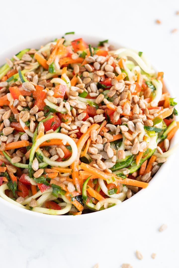 A close up of a white bowl filled with low FODMAP Asian Zoodle Salad: zucchini noodles, carrot shreds, diced red pepper, chives, cilantro and sunflower seeds tossed with an Asian-inspired vinaigrette.