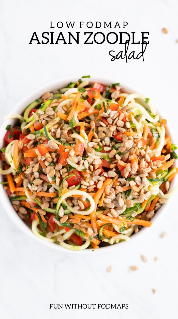 Overhead shot of a white bowl sitting on a white marble slab. The bowl is filled with low FODMAP Asian Zoodle Salad. There are random sunflower kernels artistically scattered around the bowl. In the surrounding white space, dark gray text reads low FODMAP Asian Zoodle Salad at the top and FUN WITHOUT FODMAPS at the bottom. 