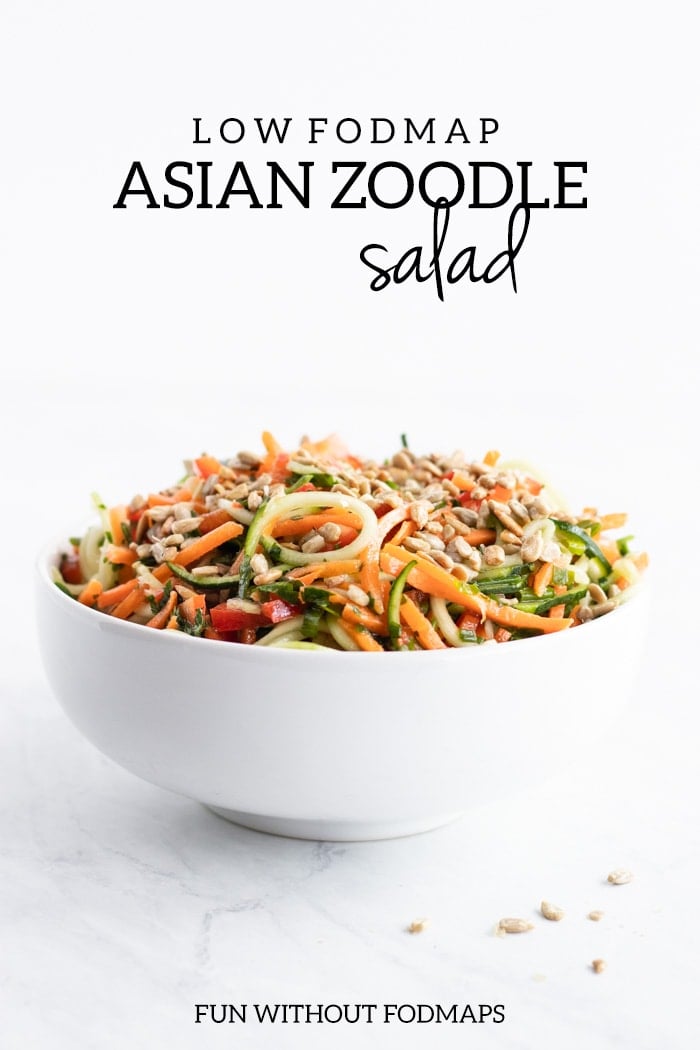 A side shot of a white bowl filled with Low FODMAP Asian Zoodle Salad. A handful of sunflower kernels are scattered in the foreground. At the top of the image dark gray text reads low FODMAP Asian Zoodle Salad.