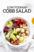 A bowl filled with lettuce, bacon, chicken, hard-boiled eggs, cherry tomatoes, and kalamata olives. Above the bowl, black text reads "Low FODMAP Cobb Salad."