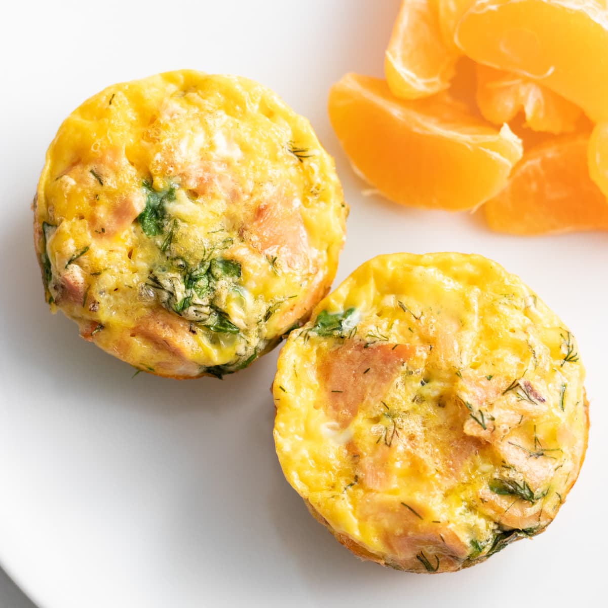 Two egg muffins made with smoked salmon, dill and spinach sit on a small white plate with orange segments.