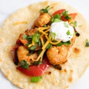 A corn tortilla topped with cooked shrimp, tender bell pepper strips, sour cream, cilantro, and shredded cheese.