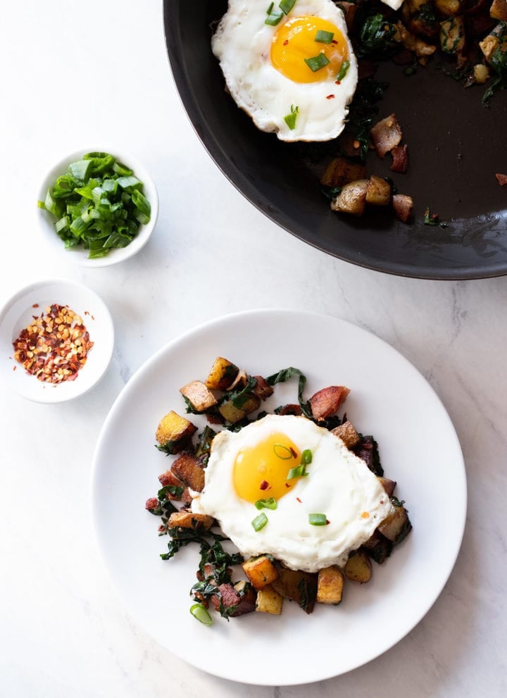 A skillet and plate containing low FODMAP Bacon, Chard, and Potato Hash sit on a white marble slab. There are also white pinch bowls filled with red pepper flakes and sliced green onion tops.