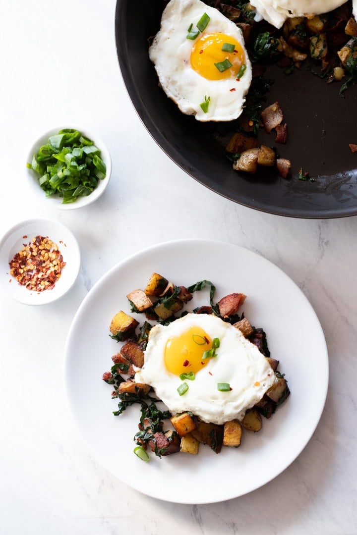 A skillet and plate containing low FODMAP Bacon, Chard, and Potato Hash sit on a white marble slab. There are also white pinch bowls filled with red pepper flakes and sliced green onion tops.