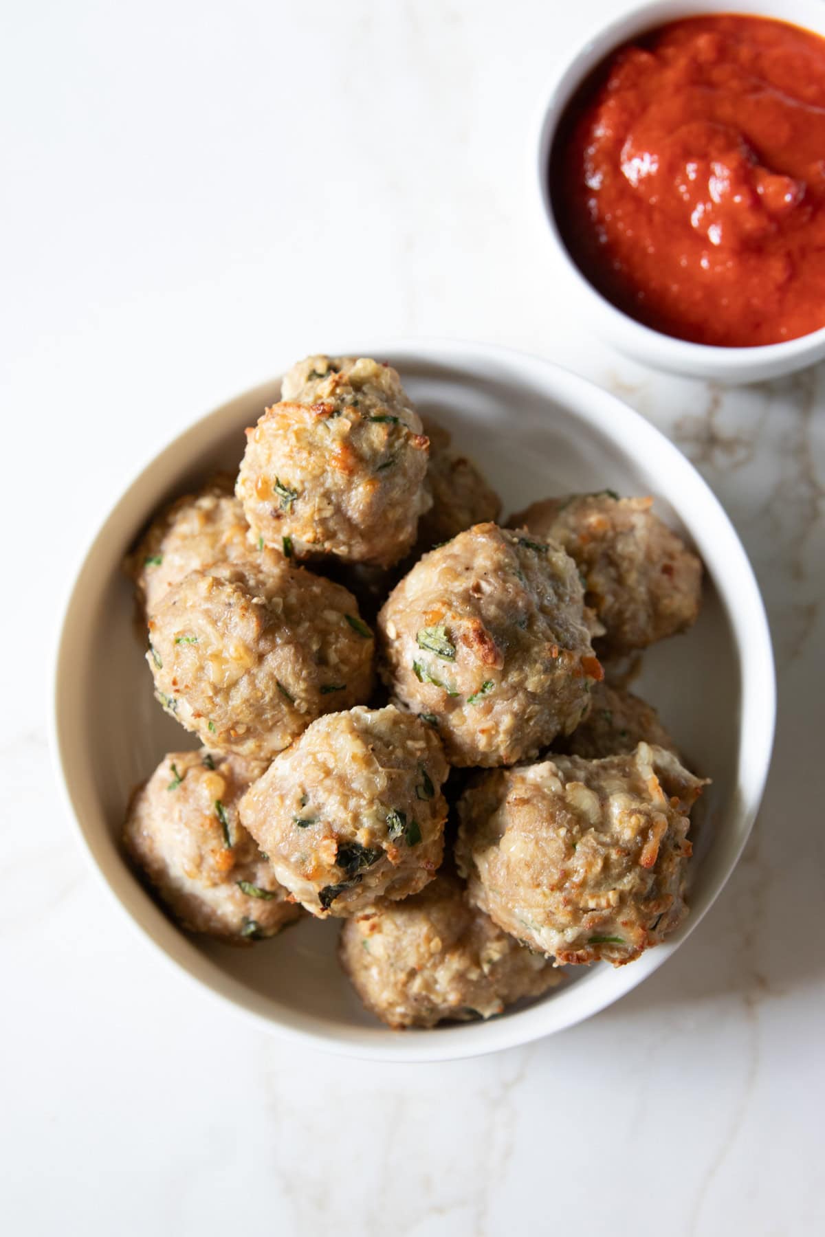 A large bowl of baked low FODMAP pesto turkey meatballs sits on a white counter with a small bowl of low FODMAP marinara off to the side.
