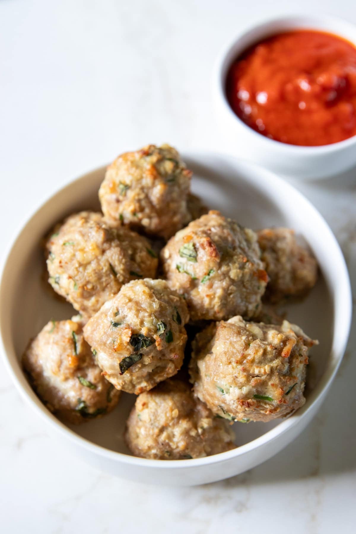 Two bowls sit on a white counter. One is filled with stacked low FODMAP pesto turkey meatballs, the other smaller bowl is filled with low FODMAP pizza sauce.