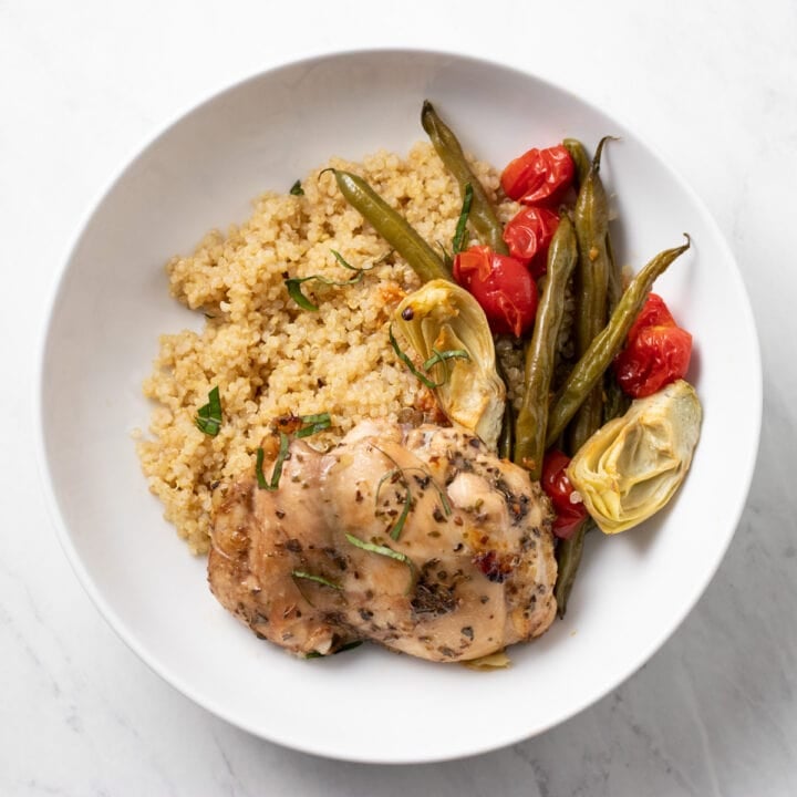 A shallow white bowl filled with cooked chicken flavored with Italian herbs, roasted green beans, cherry tomatoes, and canned artichokes, as well as cooked quinoa.