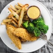 A white plate with low FODMAP chicken tenders, broccoli, fries, and a small dish of creamy maple mustard.