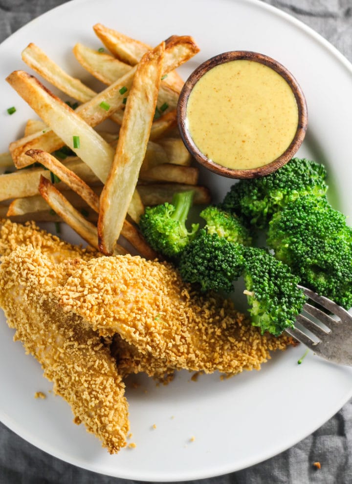 A white plate with low FODMAP chicken tenders, broccoli, fries, and a small dish of creamy maple mustard.