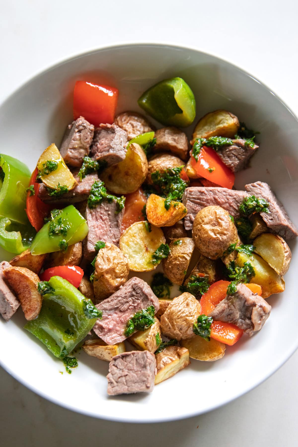 A close up of roasted potatoes, peppers and slices of top sirloin drizzled with chimichurri in a shallow bowl.
