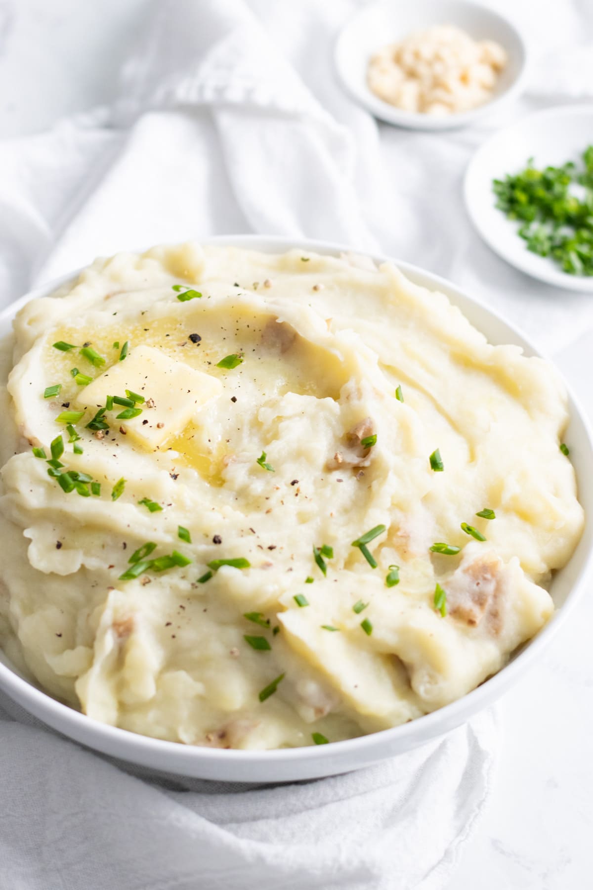 A bowl of mashed potatoes sits on a white linen napkin. The focus is on a melting dab of butter swirled into the potatoes. There are small bowls of chives and horseradish in the background.