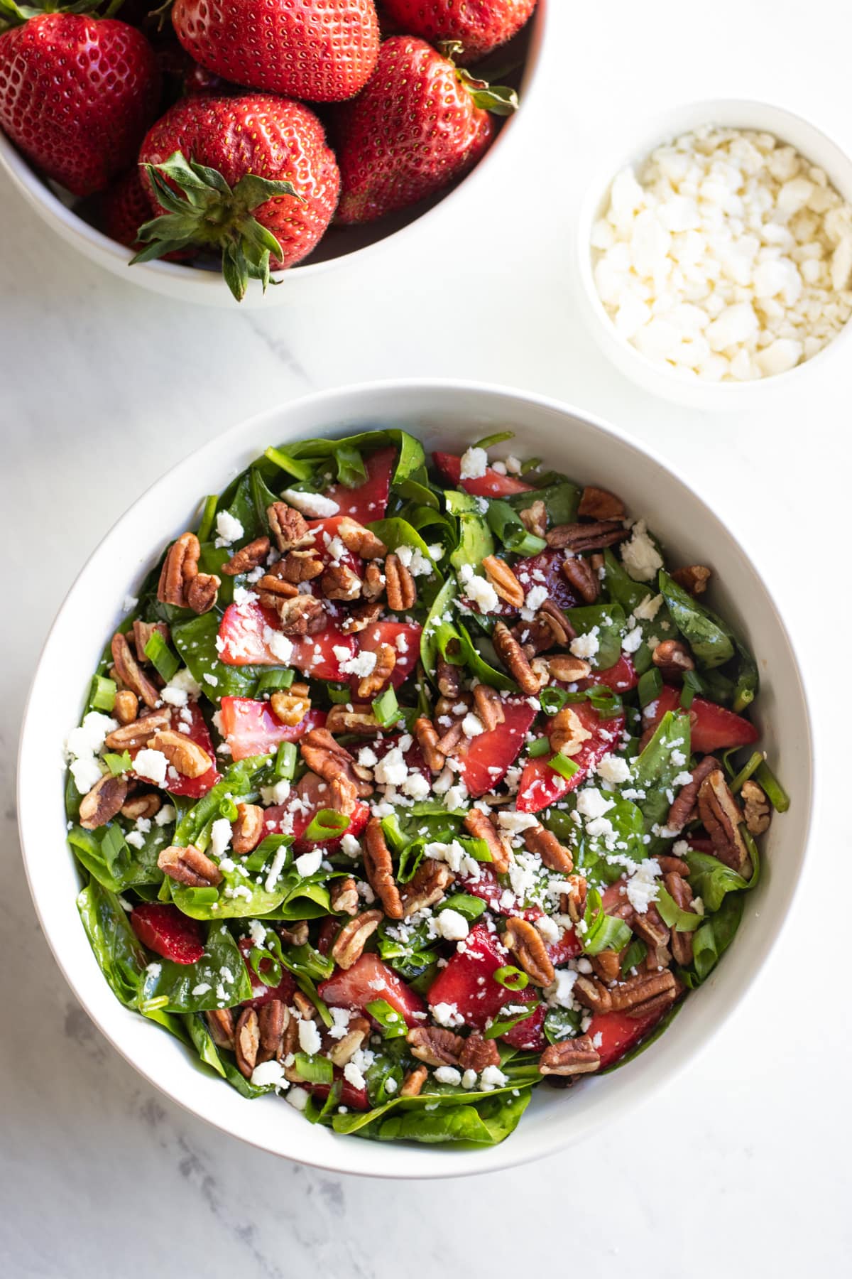 A large white bowl filled with low FODMAP spinach salad with strawberries. There are smaller bowls filled with crumbled feta cheese and whole strawberries sitting next to it.