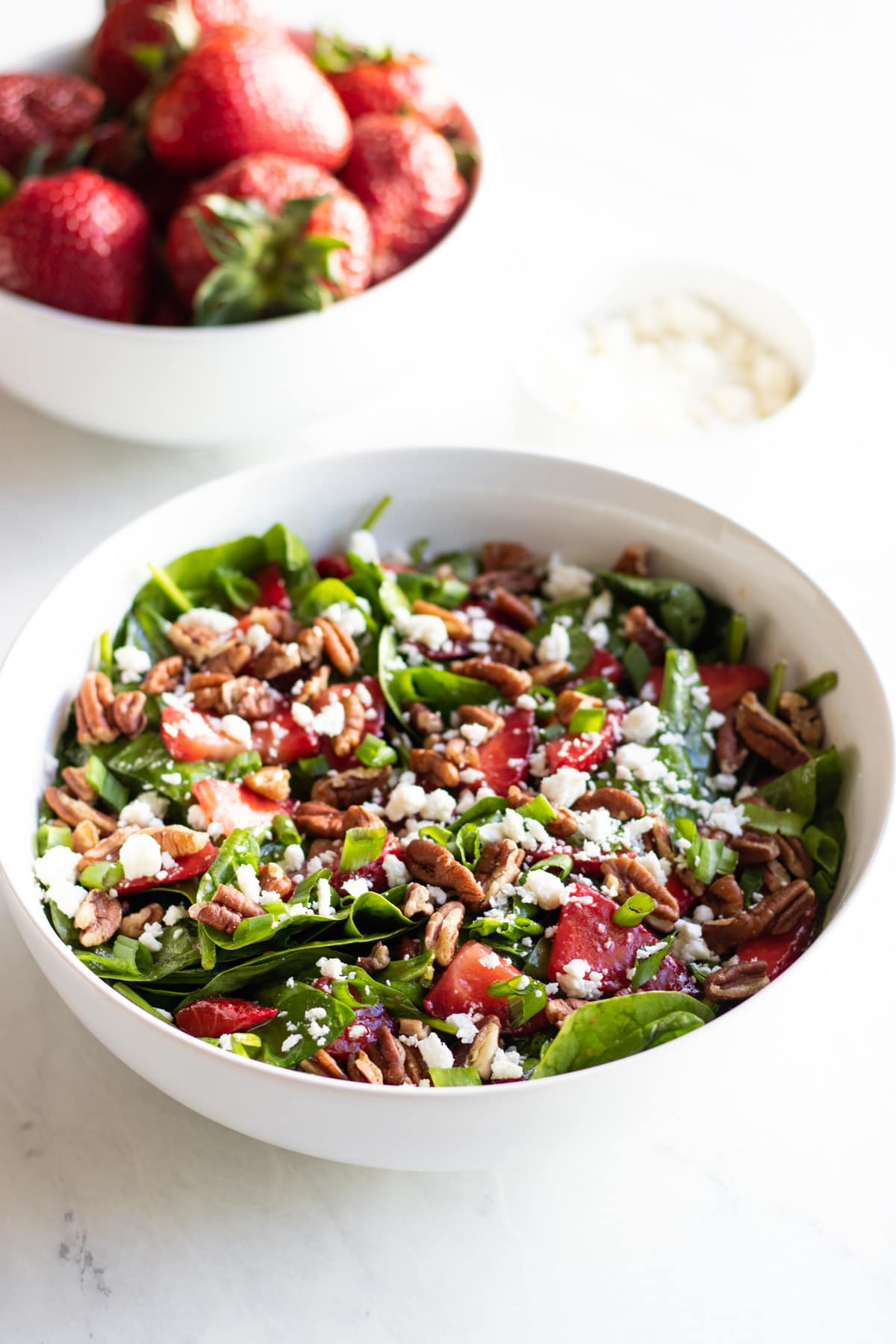 A bowl of spinach salad surrounded by smaller bowls of juicy strawberries and crumbled feta cheese.