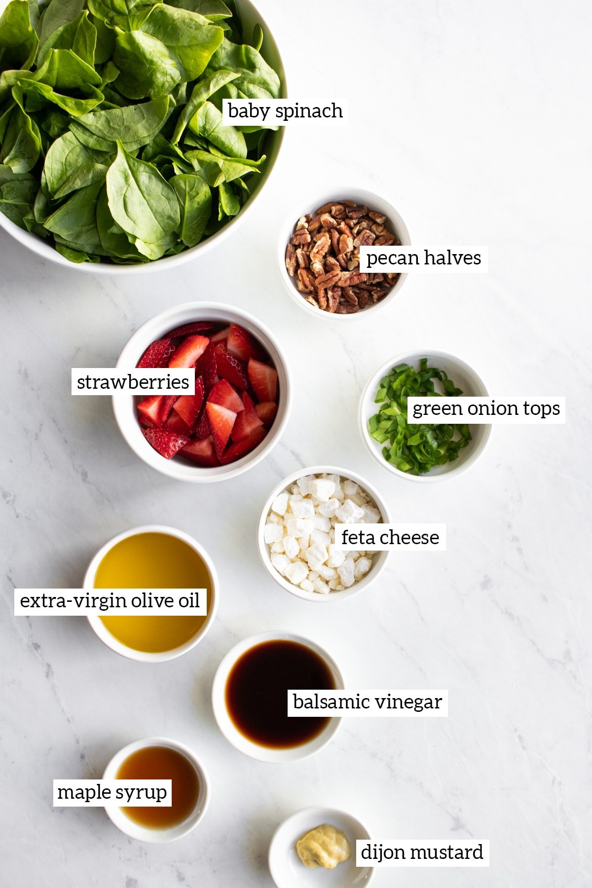 The ingredients needed for low FODMAP strawberry spinach salad are measured into small white bowls. The bowls are arranged artistically on a white marble slab. Each ingredient has a text overlay naming the food.