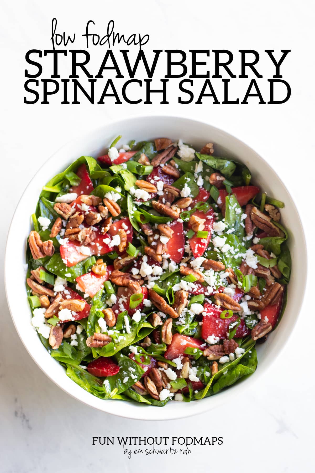 A large white bowl filled with spinach, sliced strawberries, crumbled feta, pecan pieces, and sliced green onion tops. Above the bowl black text reads "Low FODMAP Strawberry Spinach Salad". Below the bowl text says "FUN WITHOUT FODMAPS by Em Schwartz RDN".