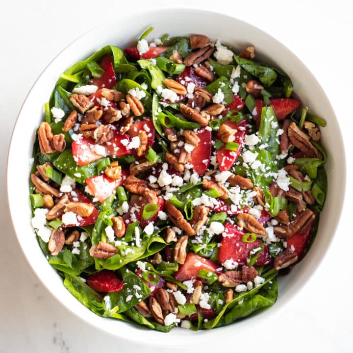 A large white bowl filled with spinach, sliced strawberries, pecans, crumbled feta cheese, and sliced green onion tops.