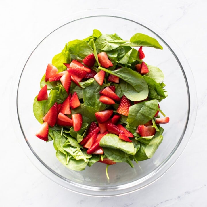 A large glass mixing bowl with spinach and sliced strawberries.