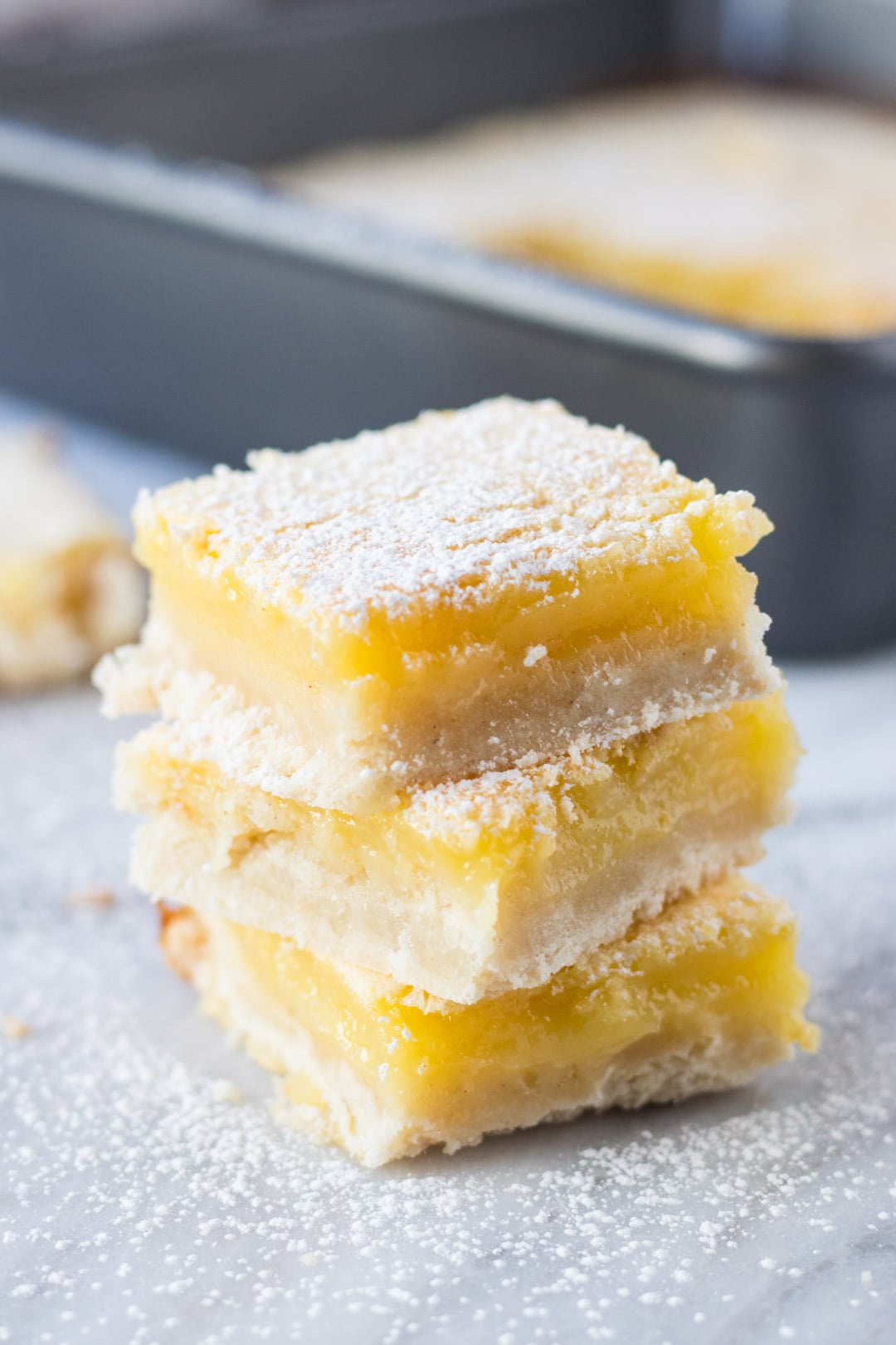 Stack of three lemon bars in front of a gray baking pan filled with remaining bars.