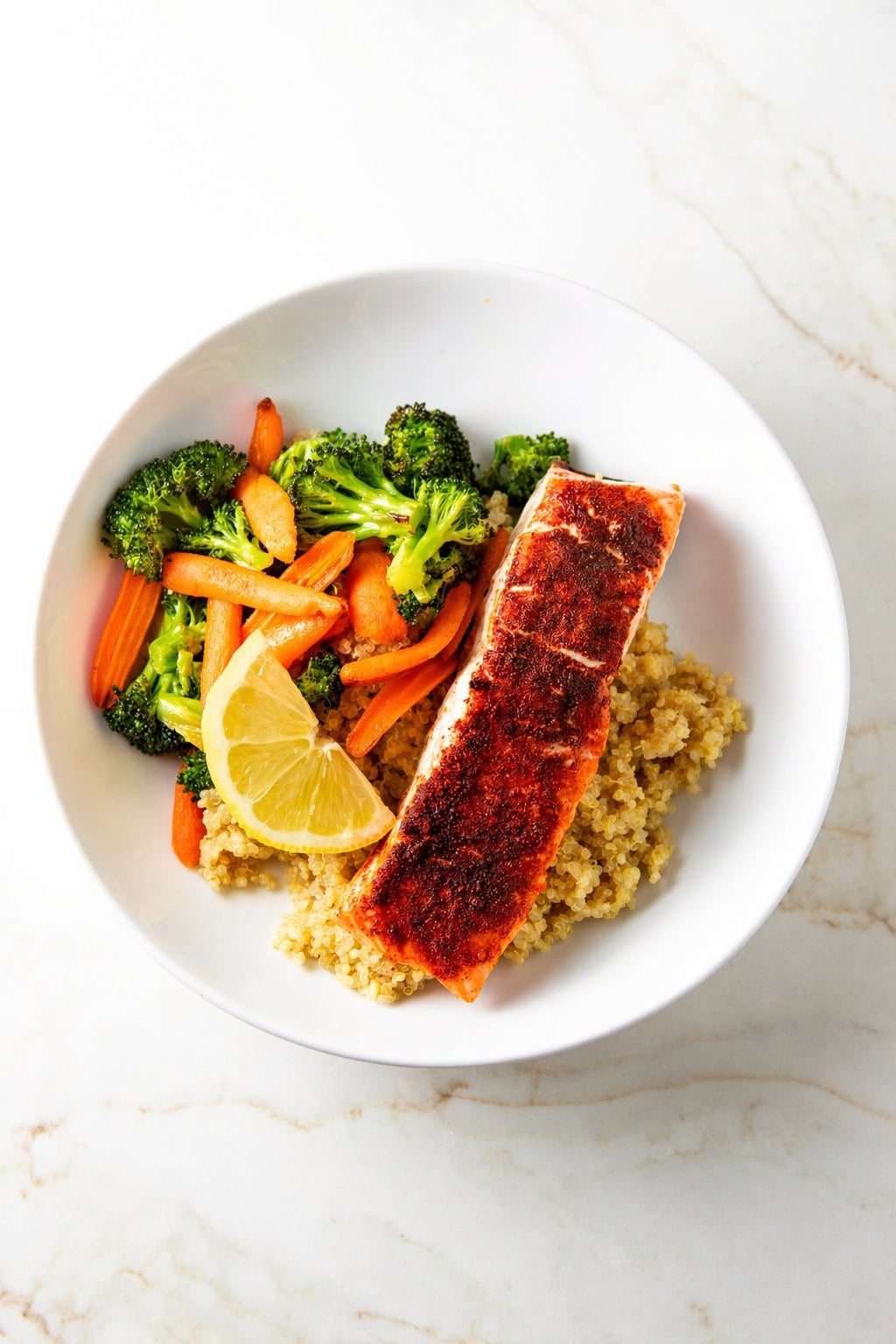 Overhead view of a white bowl with a seared salmon fillet on a bed of quinoa, served with steamed broccoli, carrot sticks, and a lemon wedge on a light marble surface.