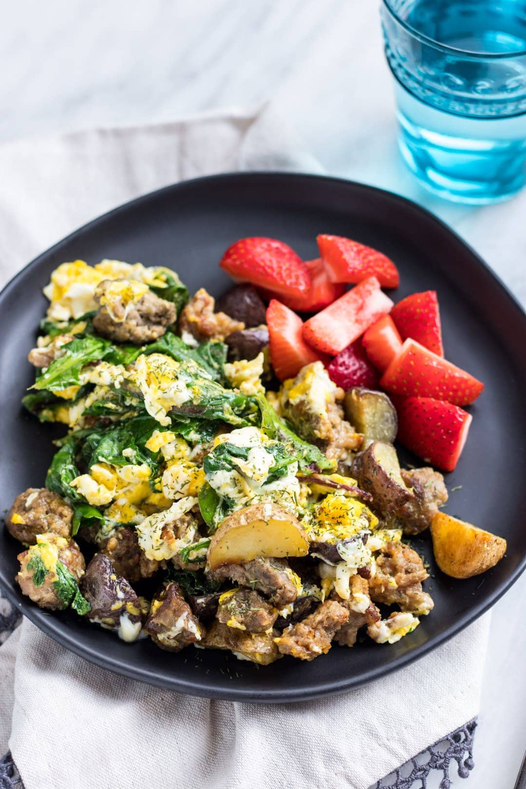 A plate of eggs scrambled with sausage crumbles, roasted potatoes, and leafy greens. There are strawberries on the side.