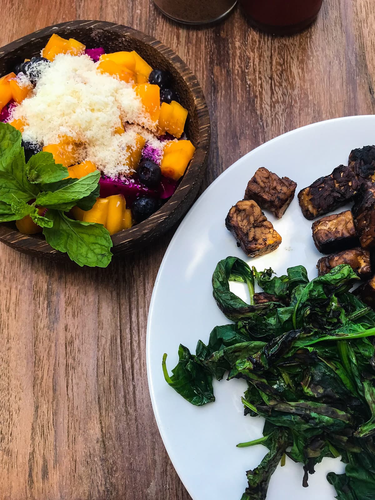 A plate of tempeh and sauteed greens with a bowl of tropical fruit on the side.