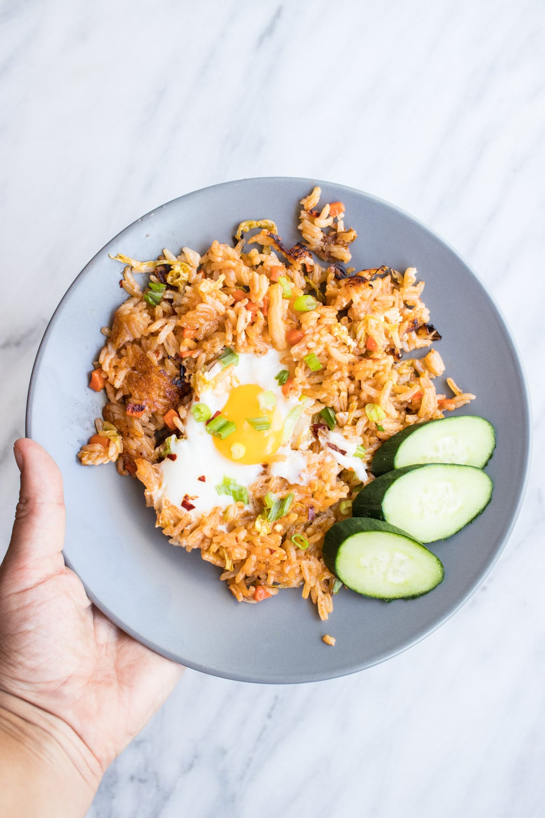 A gray plate with Indonesian fried rice topped with a fried egg and served with cucumber slices.