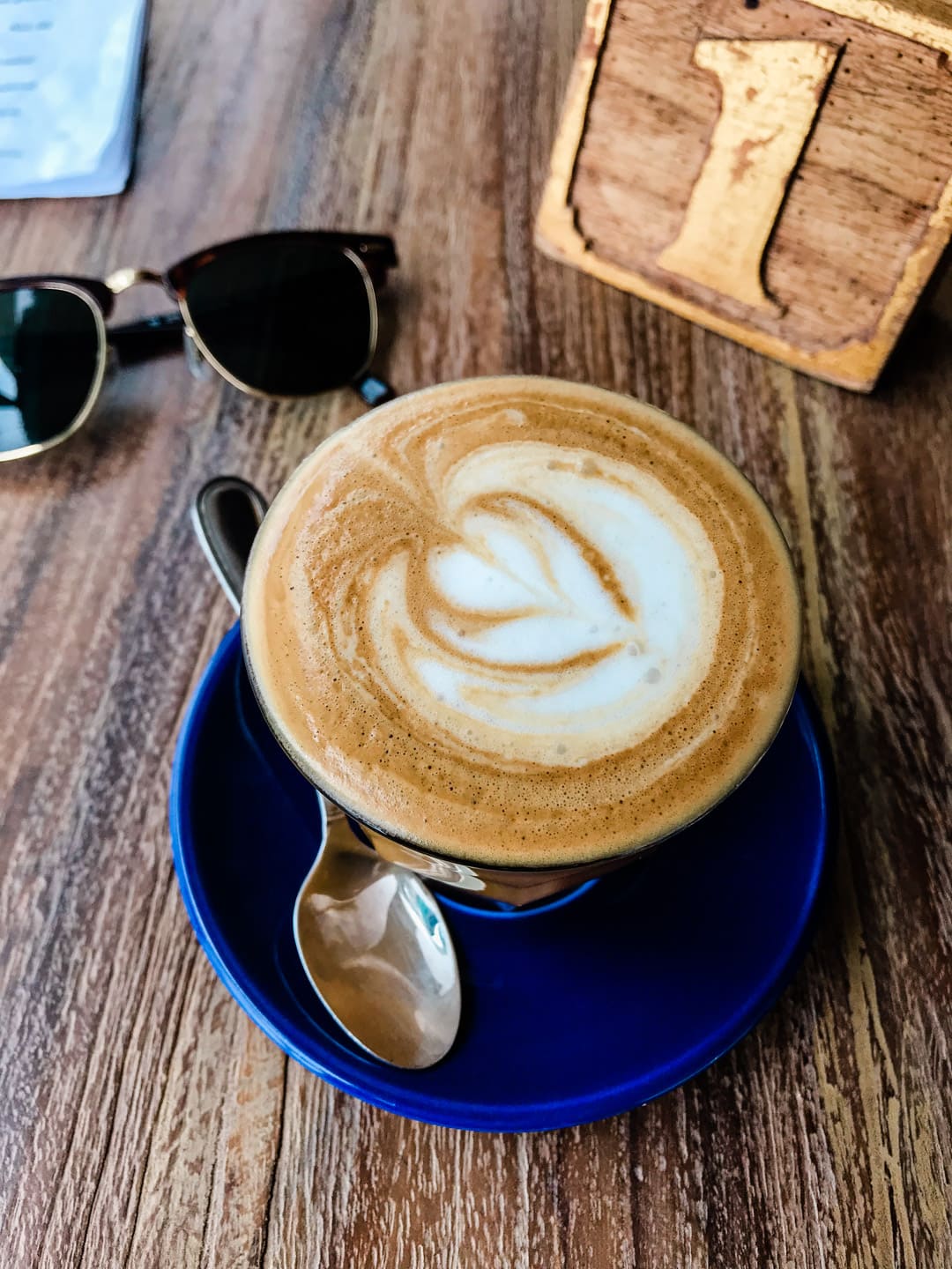 A small coconut milk latte with a small spoon on a blue plate. Rayban sunglasses and a wooden table marker with number one on it in the background.