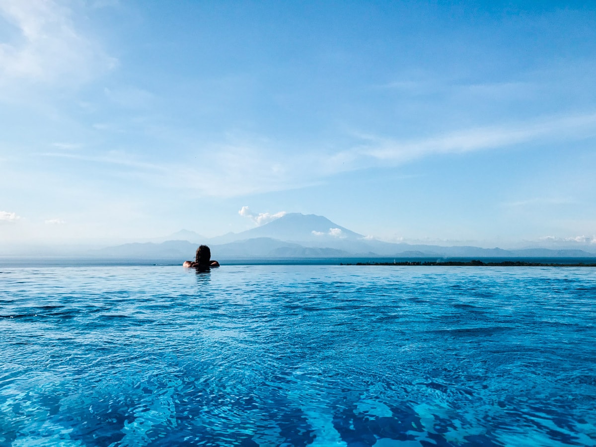 Woman swimming in an infinity pool overlooking the ocean with a volcano in the distance.