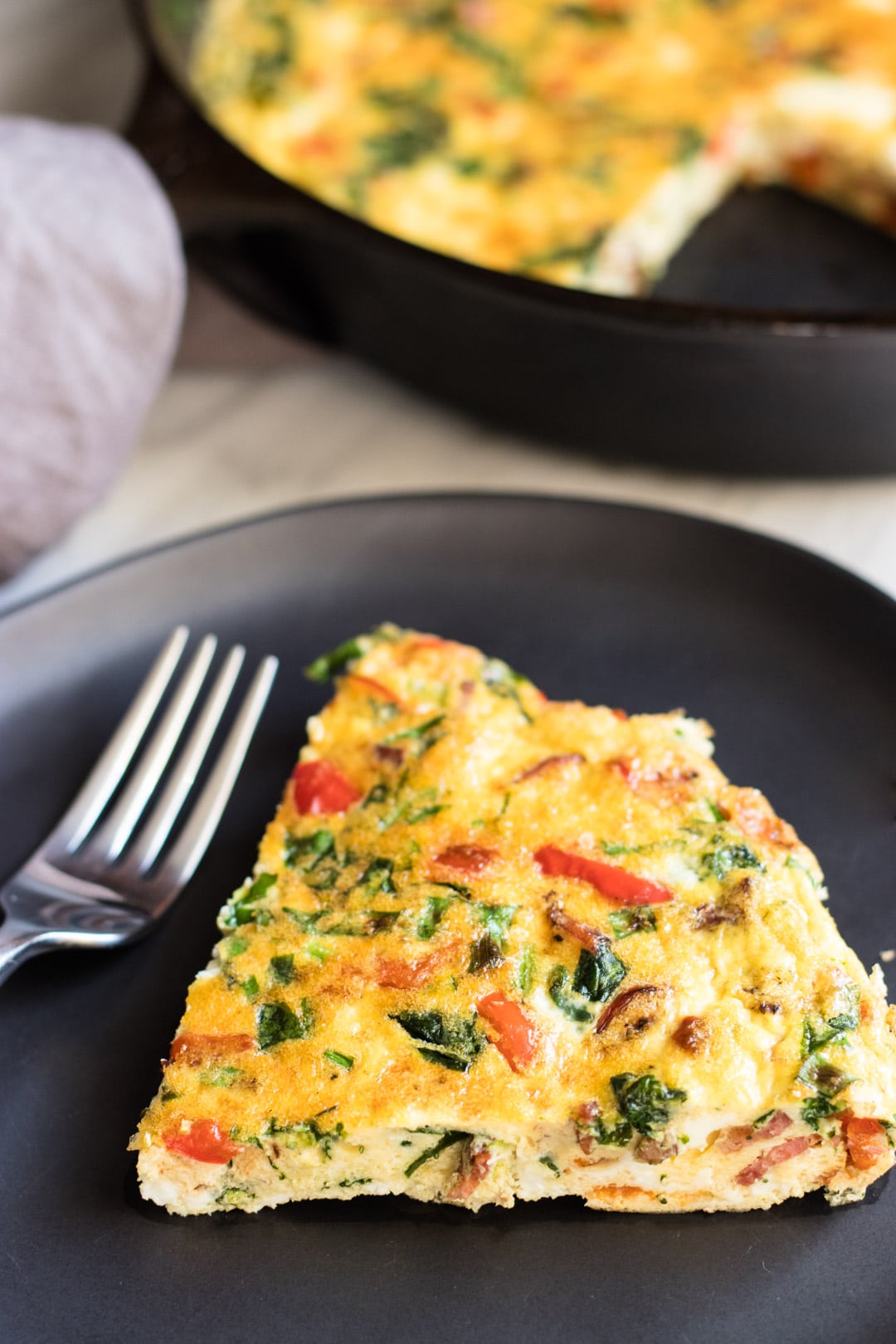 A slice of egg frittata dotted with red pepper, bacon, and green spinach sitting on a black plate with a fork.