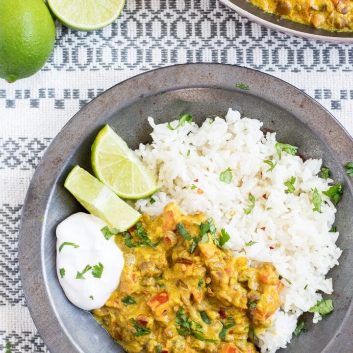 A metal bowl filled with low FODMAP lentil dal and basmati rice garnished with cilantro and served with lime wedges and a dollop of coconut yogurt. Limes and a skillet filled with dal in the background.