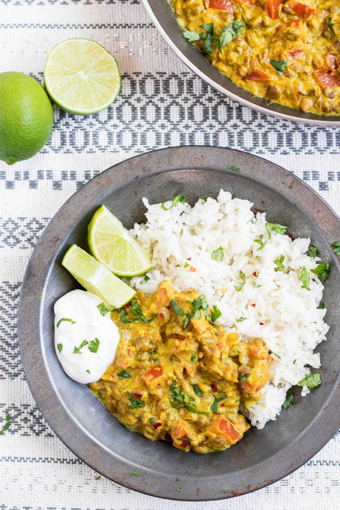 A metal bowl filled with low FODMAP lentil dal and basmati rice garnished with cilantro and served with lime wedges and a dollop of coconut yogurt. Limes and a skillet filled with dal in the background.