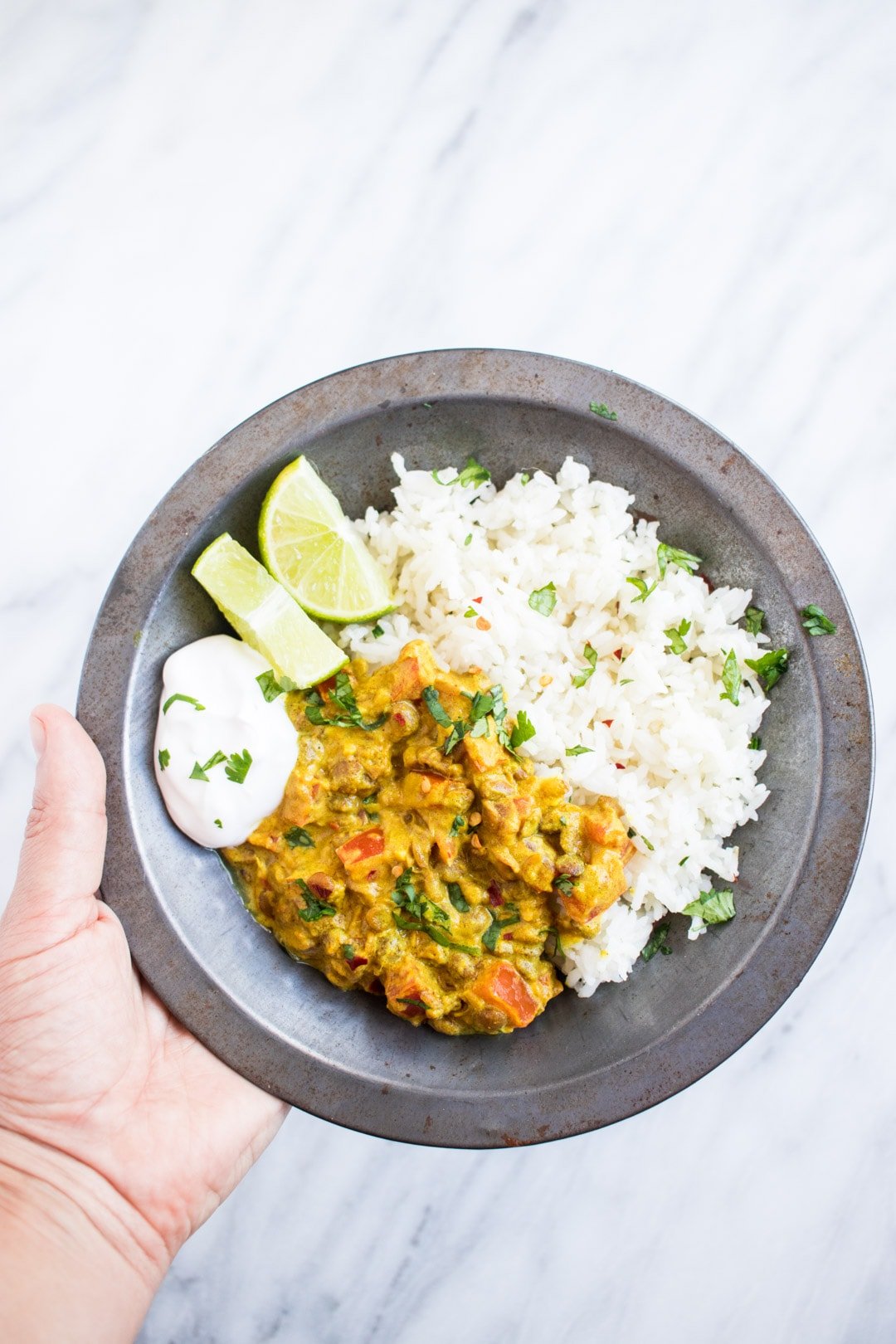 A hand holding a metal bowl filled with low FODMAP lentil dal and basmati rice garnished with cilantro and served with lime wedges and a dollop of coconut yogurt.