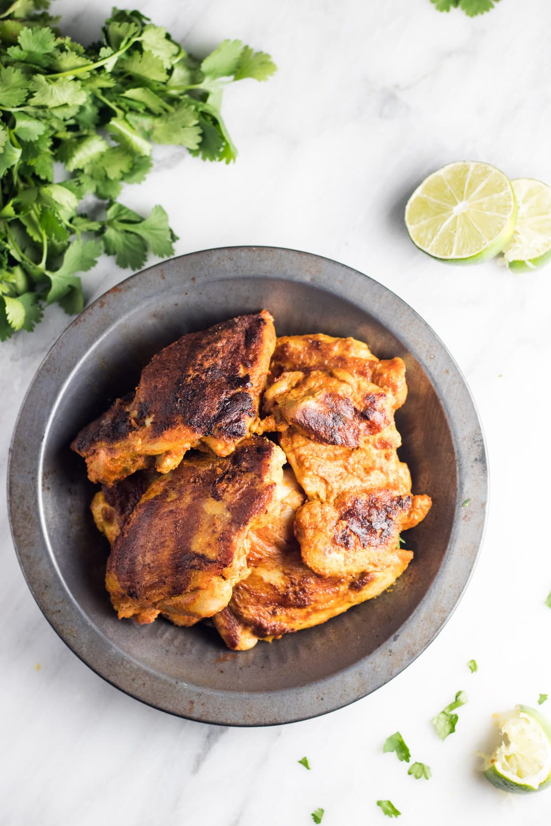 Grilled low FODMAP tandoori-inspired chicken in a metal bowl sitting on a white marble background. Cilantro leaves and lime slices surround the bowl.