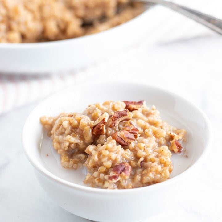 A small white bowl sits on a marble slab. It's filled with low FODMAP steel cut oats and is topped with almond milk and a couple of pecan pieces. There is a larger bowl resting in the background. It is also filled with these oats made in the Instant Pot.