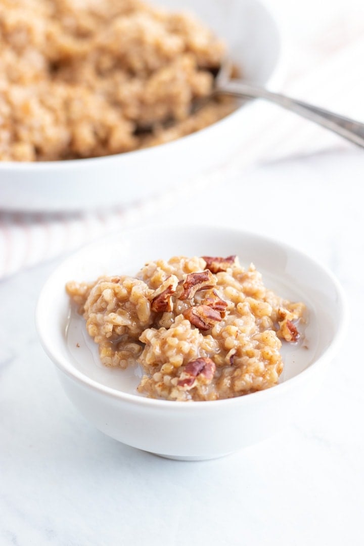 A small white bowl sits on a marble slab. It's filled with low FODMAP steel cut oats and is topped with almond milk and a couple of pecan pieces. There is a larger bowl resting in the background. It is also filled with these oats made in the Instant Pot. 